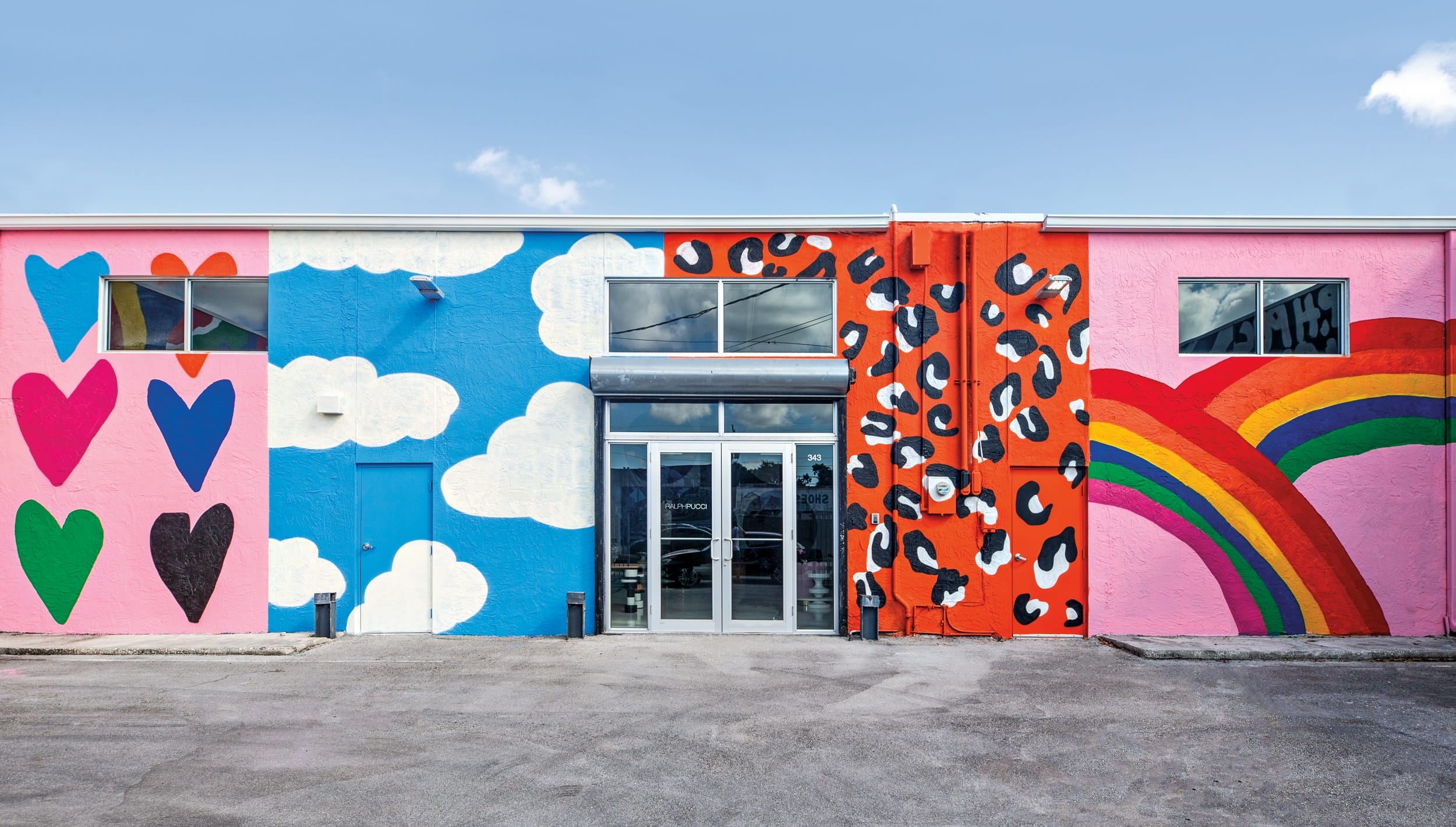 Colorful mural on building with hearts, clouds, leopard print, and a rainbow against a vibrant blue sky.