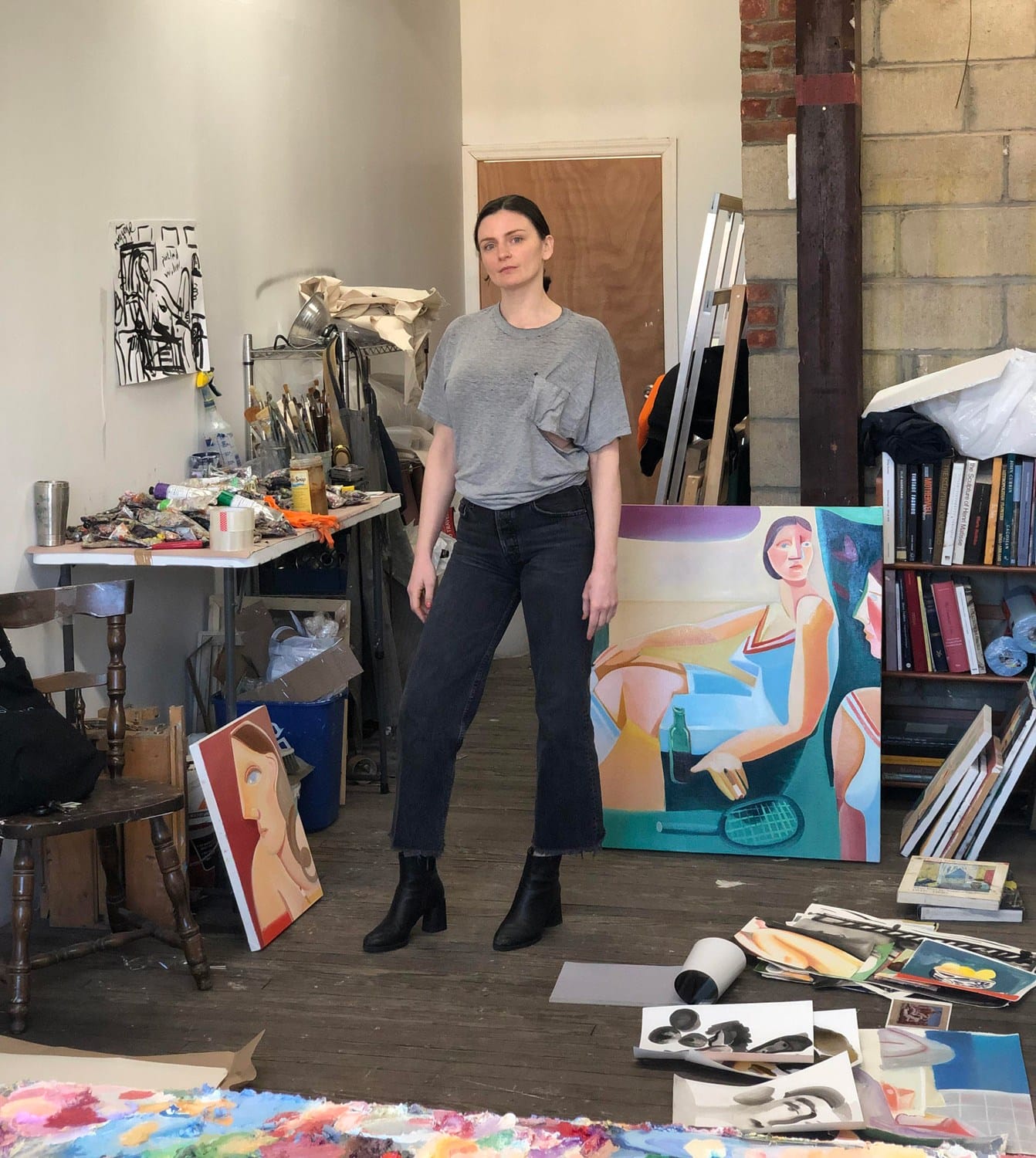 Artist standing in studio surrounded by colorful paintings and art supplies, wearing casual grey t-shirt and jeans.