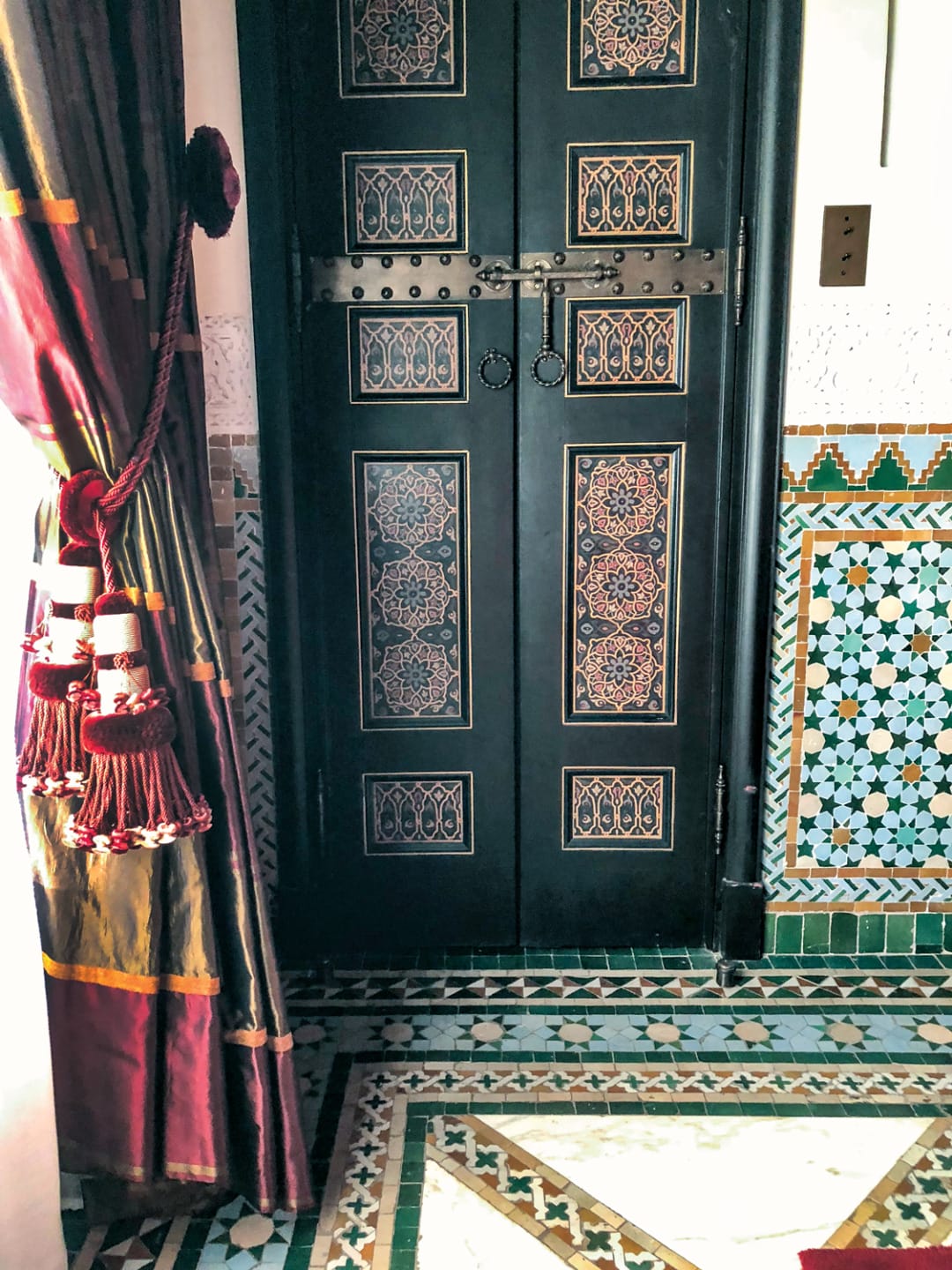 Decorative wooden door with intricate patterns, colorful mosaic tiles on walls and floor, and rich red curtain with tassels.