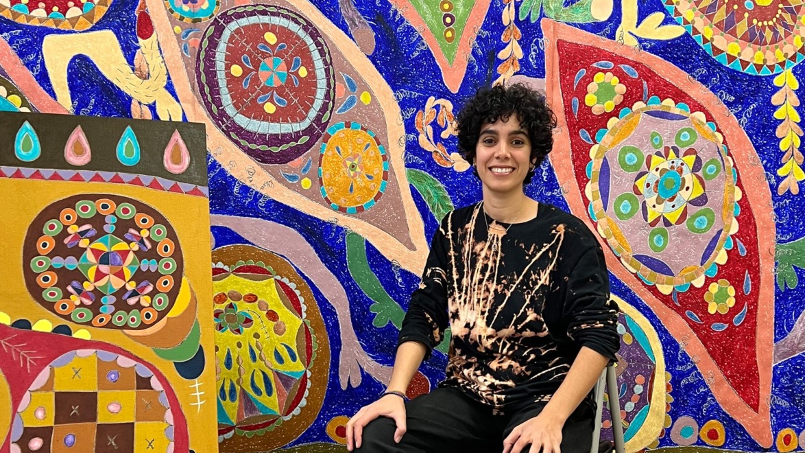 Artist seated in front of a vibrant, colorful abstract mural and painting, featuring intricate geometric designs.