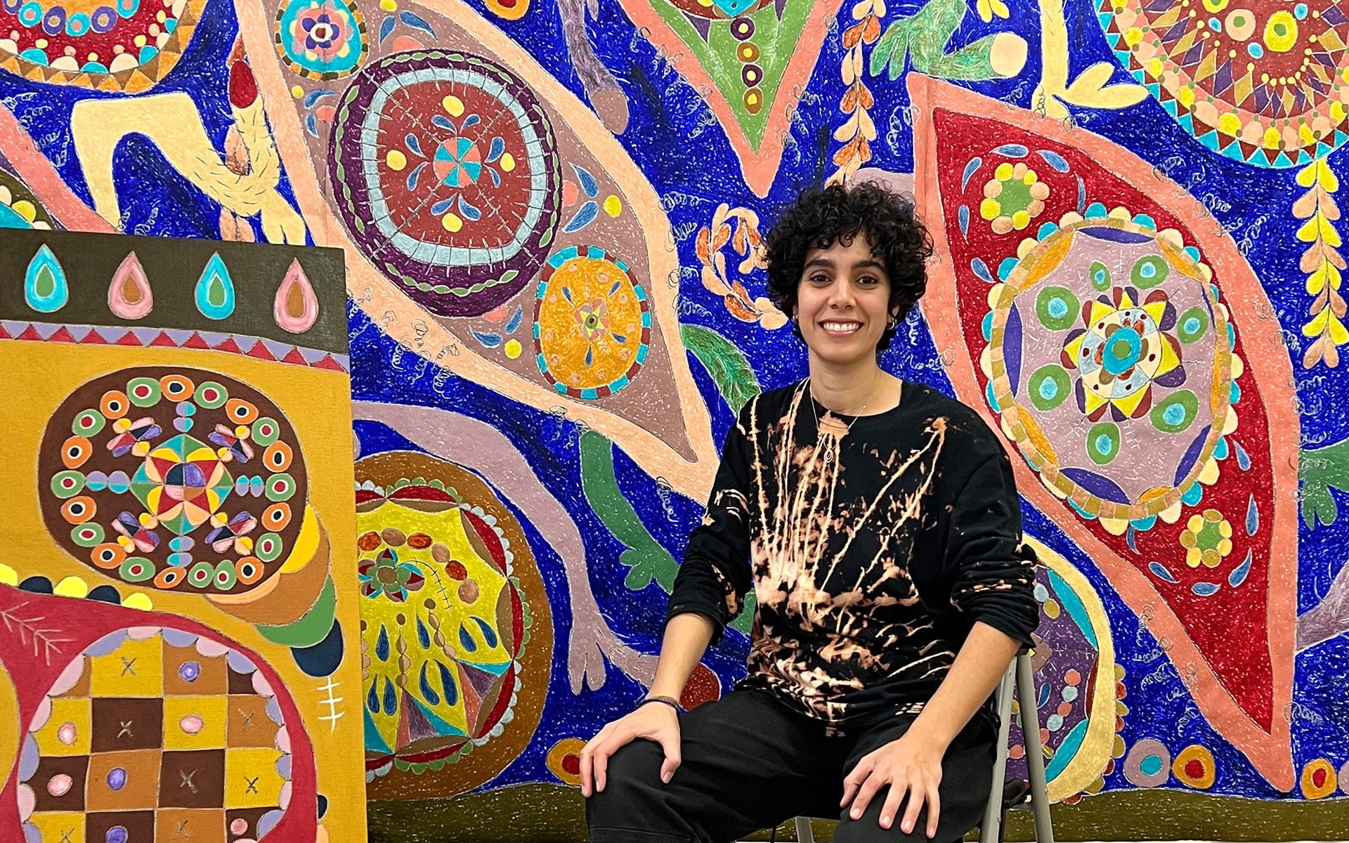 Artist seated in front of a vibrant, colorful abstract mural and painting, featuring intricate geometric designs.
