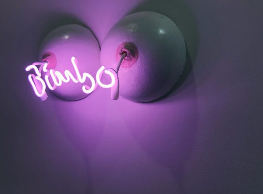 Neon sign saying "bimbo" in cursive with pink lighting against a purple wall.