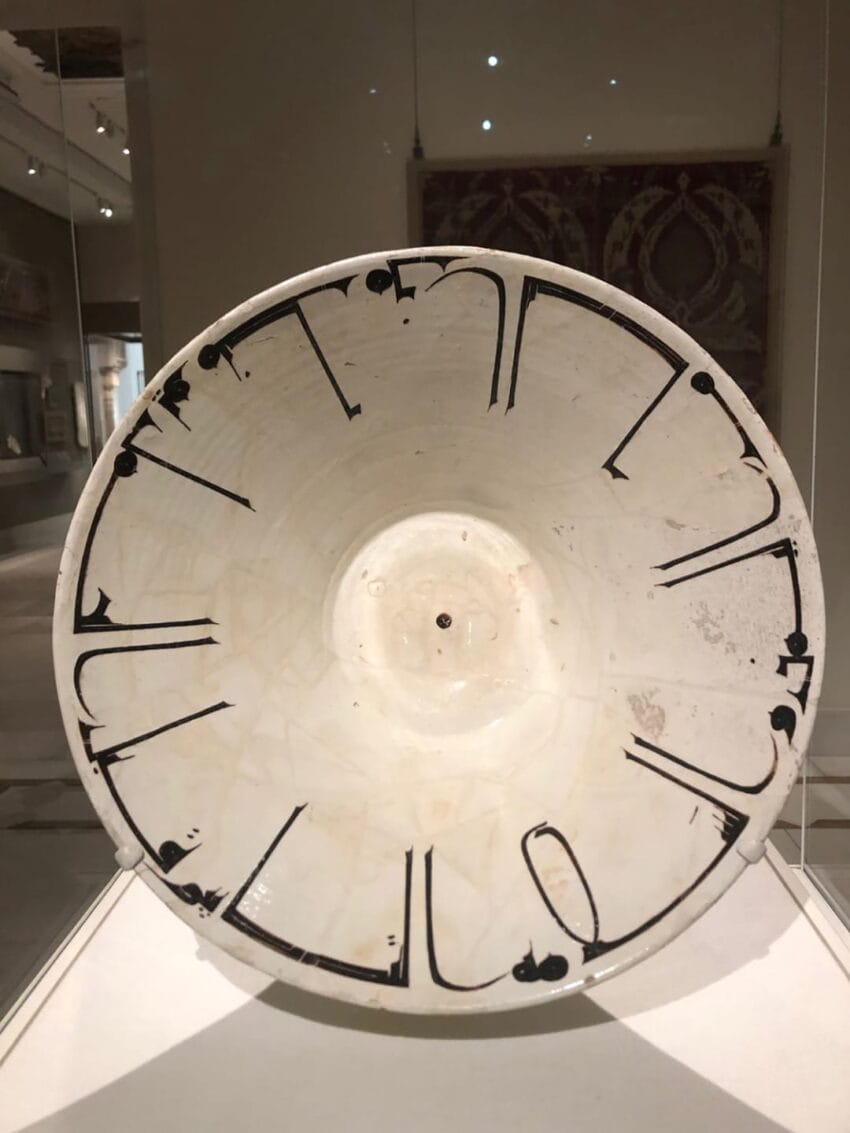 Large white ceramic bowl with black Kufic script, displayed in a museum setting.