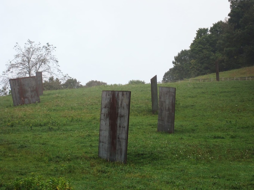 Metal sculptures standing in a grassy hillside with trees in the background on a cloudy day.