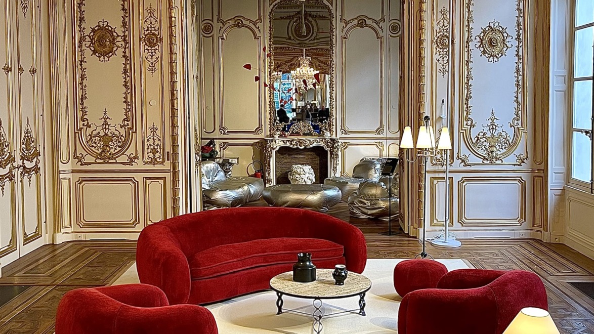 Elegant room with ornate walls, large mirror, chandelier, and modern red furniture set on a plush white rug.