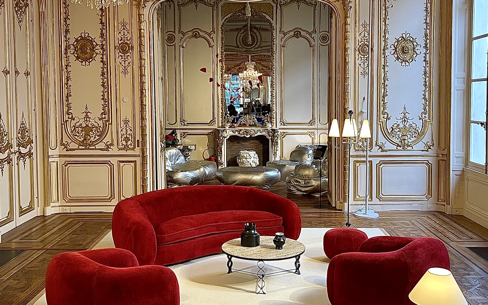 Elegant room with ornate walls, large mirror, chandelier, and modern red furniture set on a plush white rug.
