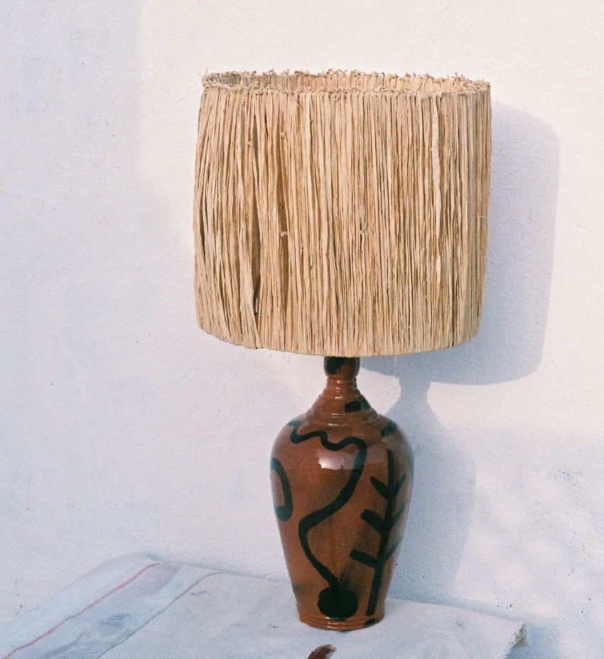Ceramic vase table lamp with a fringed straw shade on a light surface against a white wall.