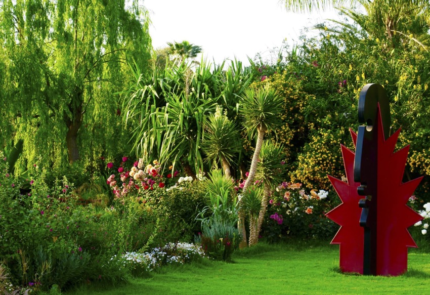 Garden with lush greenery, colorful flowers, and a modern black and red abstract sculpture on a grassy lawn.