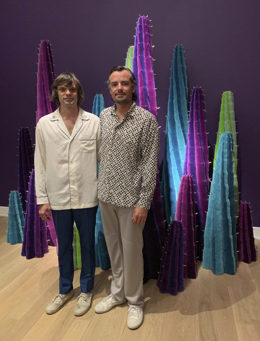 Two people standing in front of colorful art installation with tall, textured, cone-like structures on wooden floor.