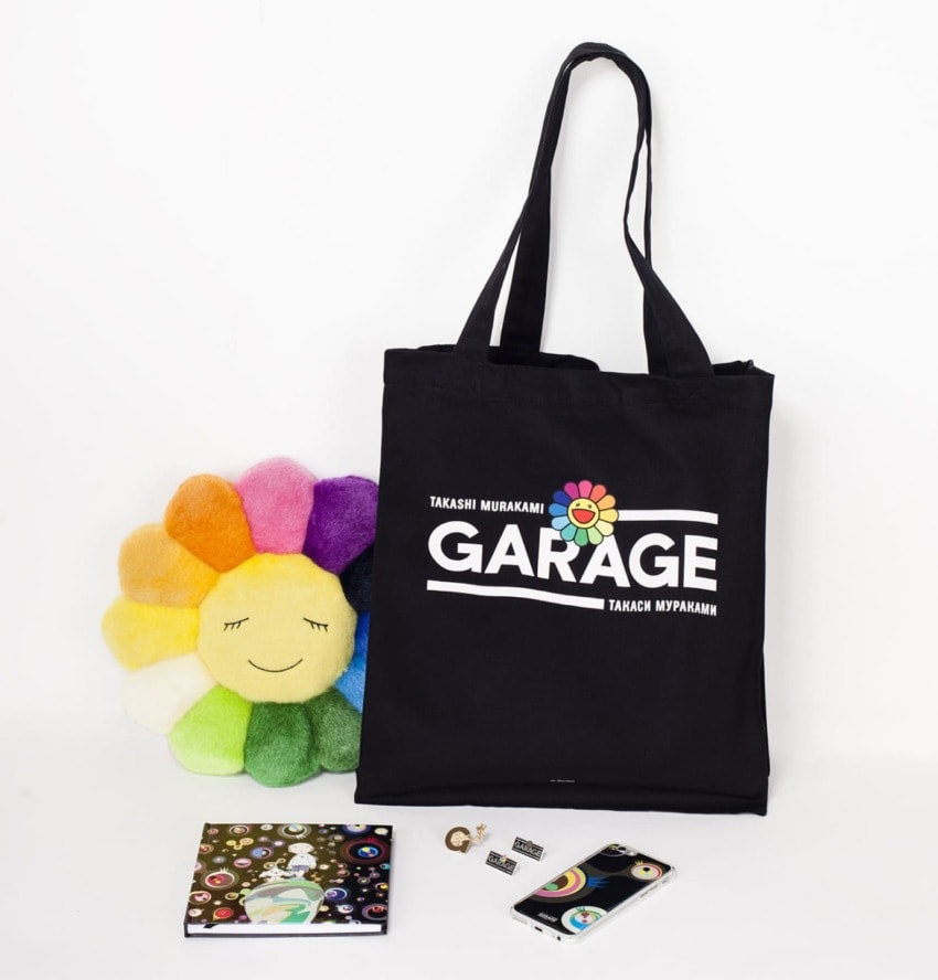 Black tote bag with colorful smiling flower, notebook, keychain, and phone case displayed on white background.
