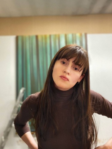 Person with long brown hair and bangs wearing a dark top stands with hands on hips in a room with green curtains.