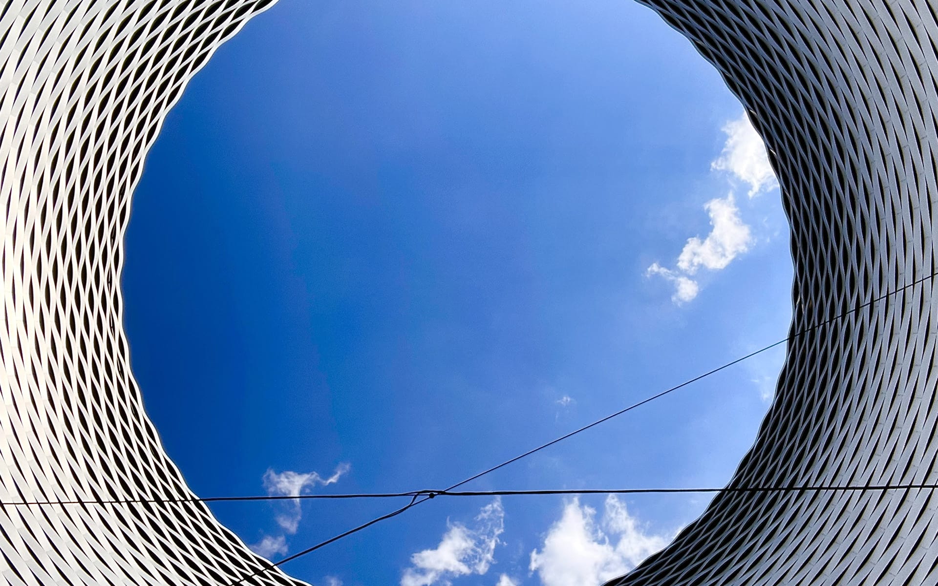 View of a cloudy blue sky through a circular architectural opening with a modern, textured design and intersecting cables.