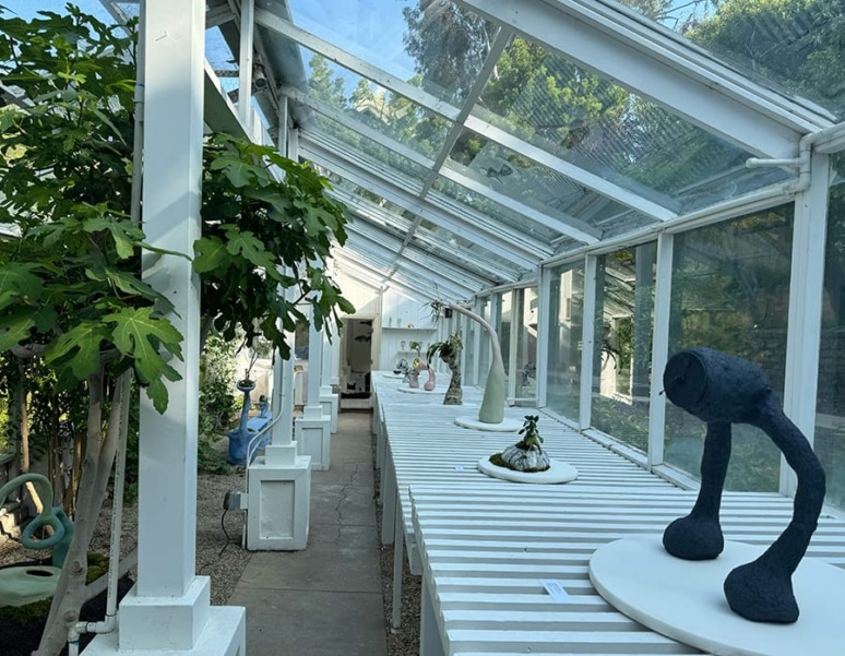 Sculptures displayed on white tables inside a sunlit greenhouse with lush greenery in the background