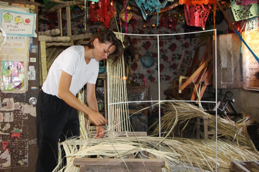 Person weaving straw in a colorful, decorated workshop with handmade crafts and vibrant hanging decorations.