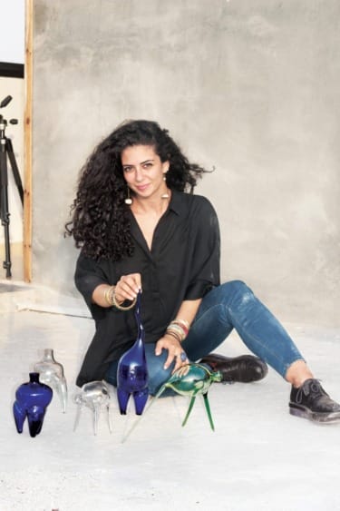 Person sitting on floor with glass sculptures, wearing black shirt and jeans, in front of a concrete wall with a tripod nearby.