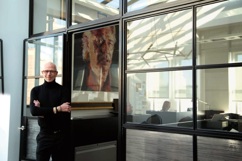 Man in black sweater standing by a glass office door with a colorful portrait in the background.