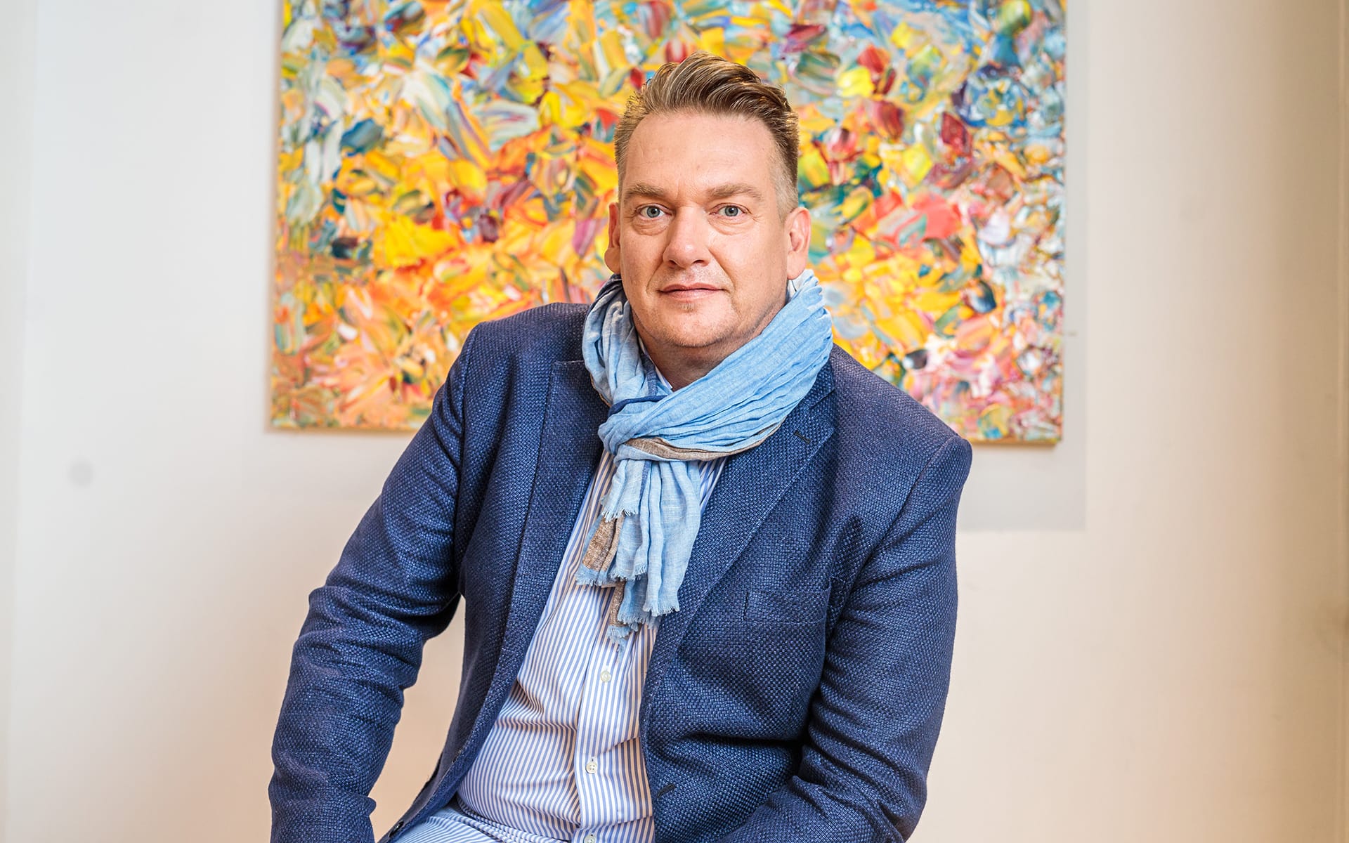 Man in blue blazer and scarf sitting in front of a colorful abstract painting in a bright room.