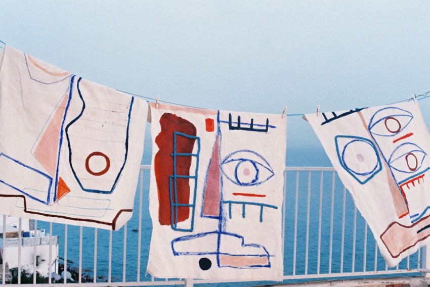 Abstract painted canvases hanging on a clothesline with a sea view in the background.
