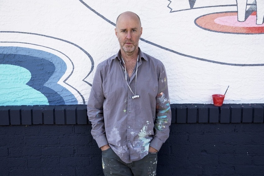 Man with paint-splattered shirt standing against a mural-covered wall, holding painting tools.