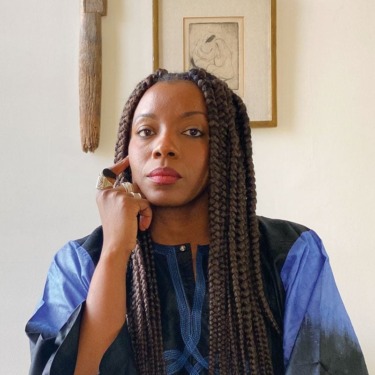Person sitting thoughtfully with long braided hair, wearing a blue and black outfit, with art in the background.