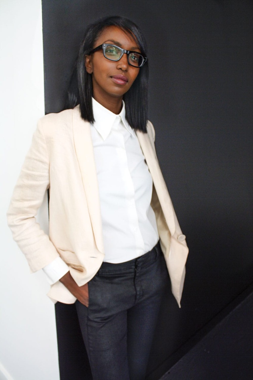 Person wearing a beige blazer and white shirt, standing against a black wall, looking confidently at the camera.