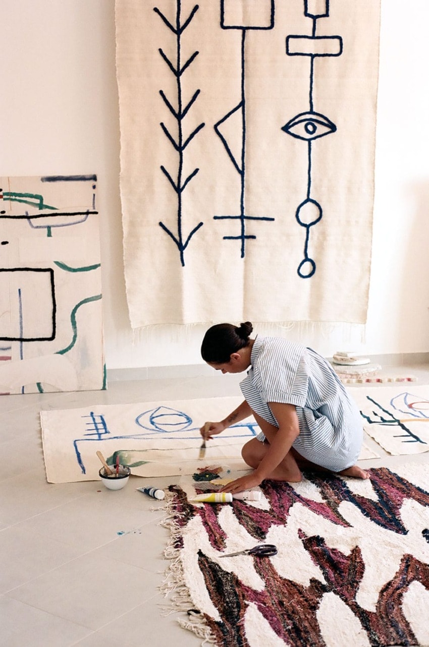 Artist painting abstract designs on canvas rugs on the floor, with other artistic rugs hanging on the wall.