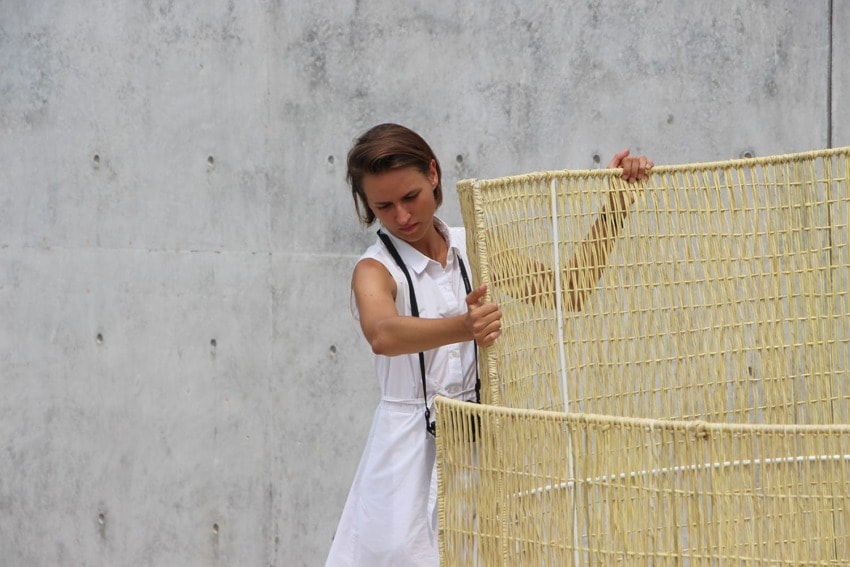 Person in a white dress adjusting a woven structure against a concrete wall backdrop.
