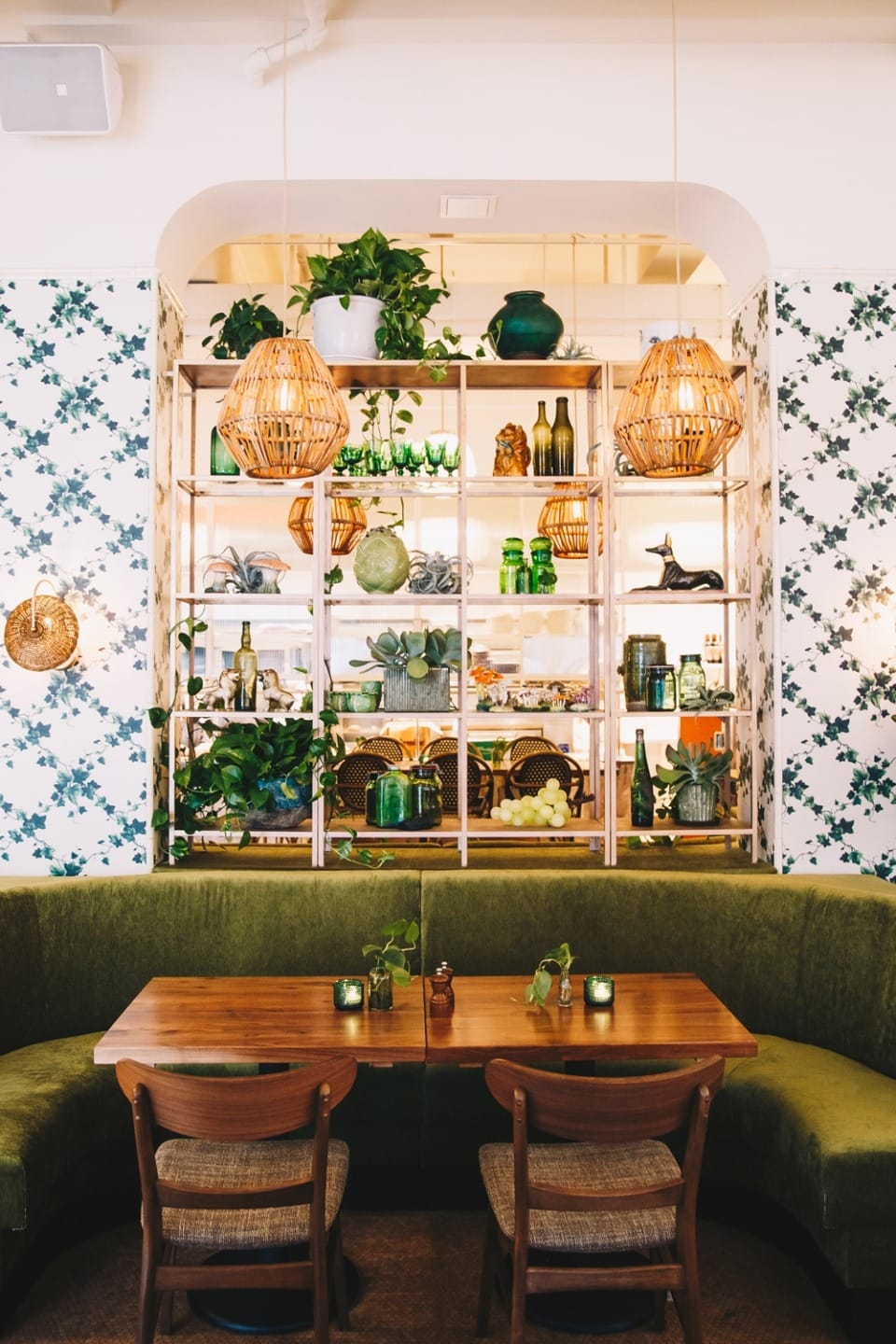 Cozy cafe interior with wooden table, green velvet seating, hanging wicker lights, and floral wallpaper.
