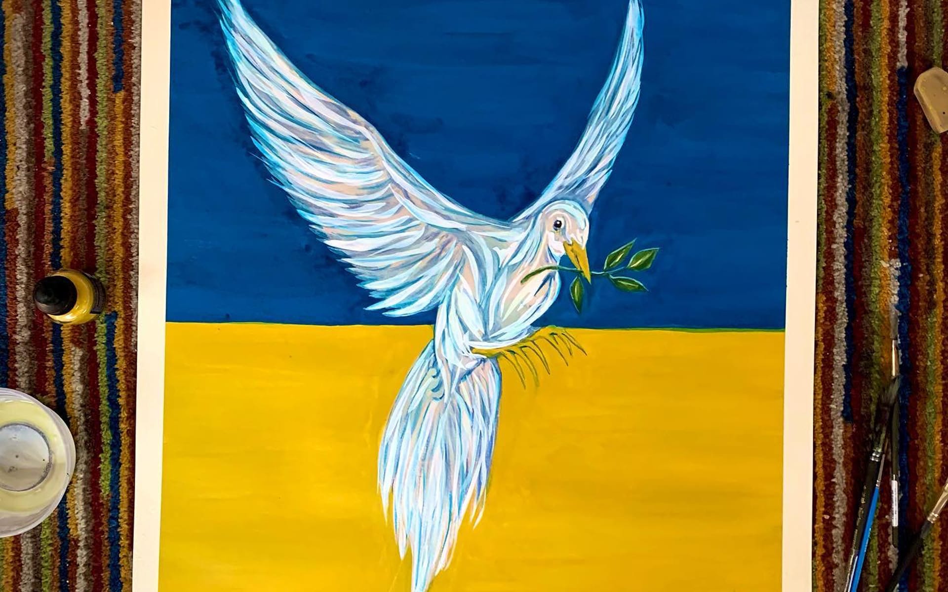 Painting of a white dove holding an olive branch, set against a blue and yellow background, surrounded by art supplies.