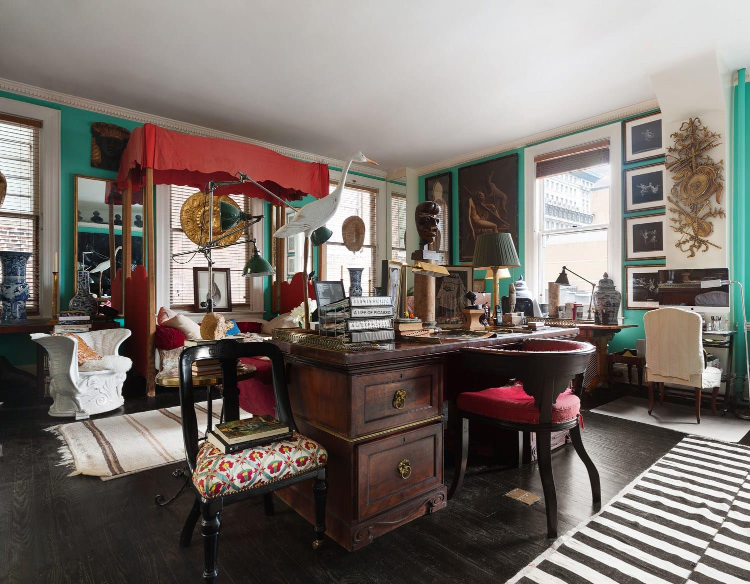 Eclectic home office with vintage wooden desk, red canopy bed, and vibrant decor in a room with dark hardwood flooring.