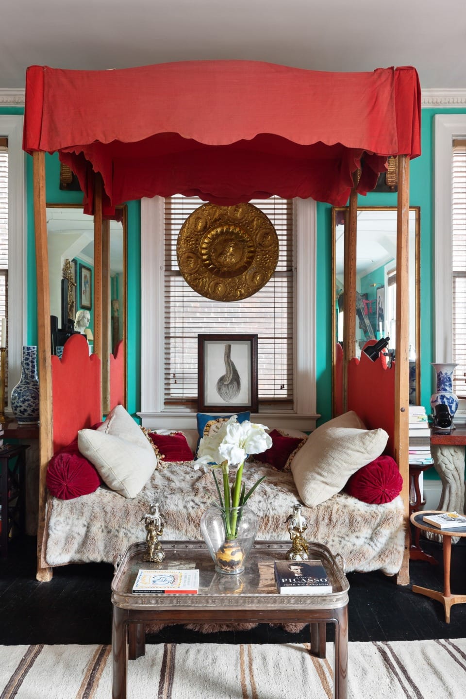 Red canopy bed in vibrant room with teal walls, gold wall art, and coffee table with flowers and decorative items.