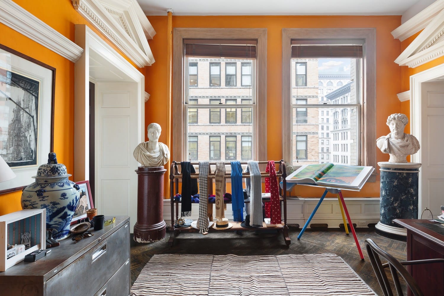 Colorful room with busts, city view windows, and art display. Decor includes vases, books, and scarves on a rack.