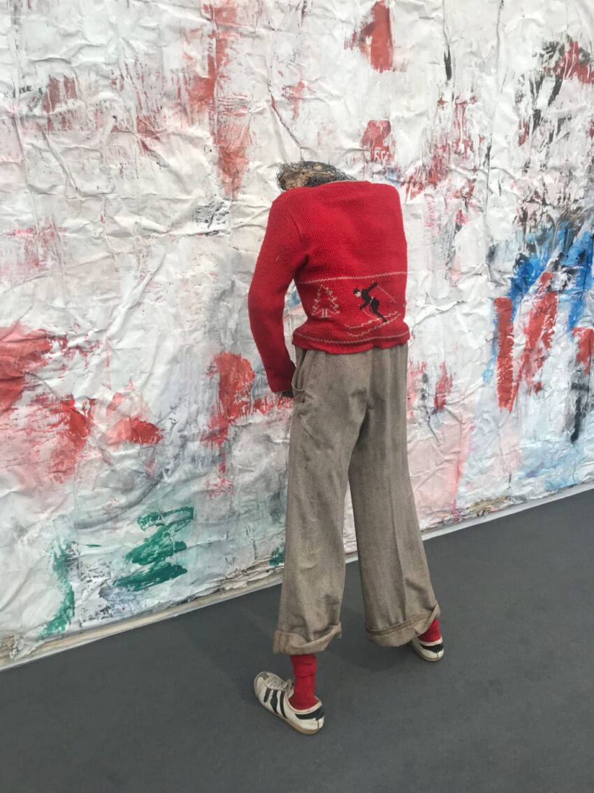 Abstract art installation of a headless mannequin wearing a red sweater and gray pants, set against a colorful textured backdrop.