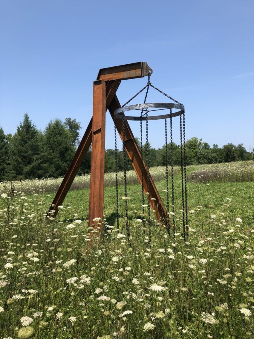 Rustic metal sculpture in a field of wildflowers with trees in the background and a clear blue sky overhead.