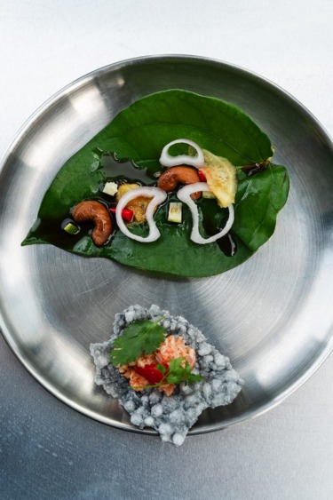 Elegant dish with leaf, nuts, and vegetables on a metal plate, accompanied by a small, garnished clump of rice.