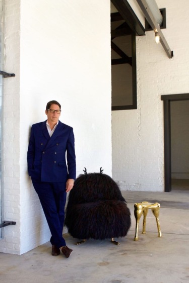 Man in blue suit leans against a white wall beside a unique modern chair and gold stool in a spacious industrial room.