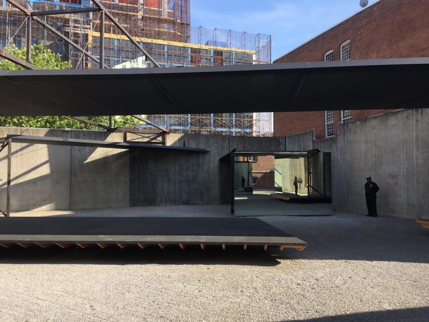 Outdoor art installation with large black platforms and mirrors in a concrete courtyard, surrounded by brick and industrial buildings.