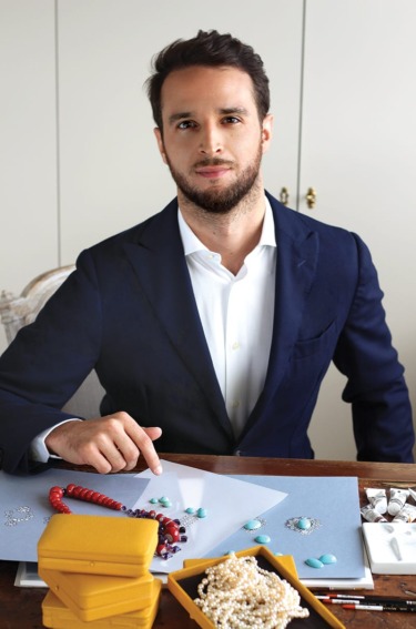 Man in a suit sitting at a table with jewelry designs and boxes, pointing at colored gemstones on a paper.