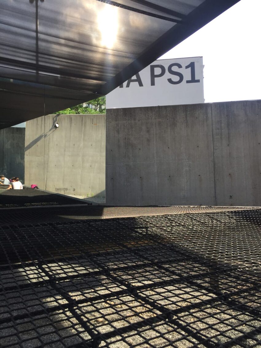 View of a contemporary art museum entrance with shadows cast on the ground and a concrete wall in the background.