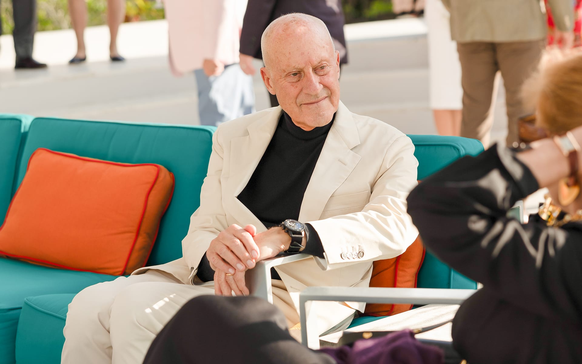 Elderly man in a cream suit sitting on a turquoise sofa with orange pillows, conversing with a person in a black outfit.