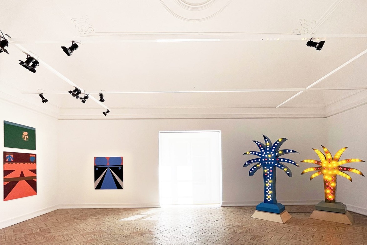 Art gallery with colorful geometric paintings on walls and two illuminated palm tree sculptures on wooden floor.