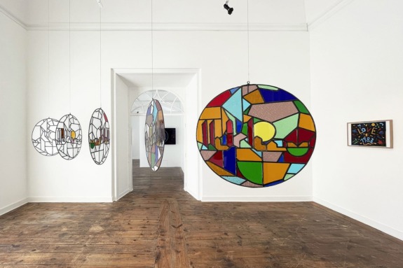 Colorful stained glass art pieces hanging in a gallery with wooden flooring and white walls.
