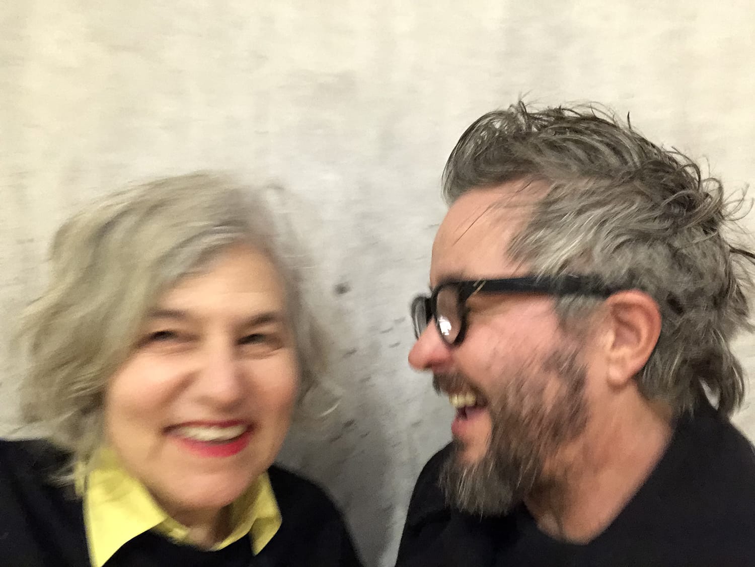 Two people smiling and laughing together in a blurred photo, one with gray hair and the other with a beard and glasses.