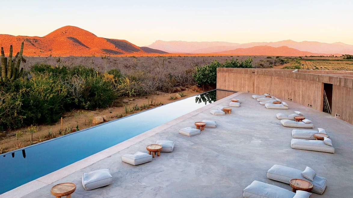 Desert landscape with a modern outdoor seating area and infinity pool at sunset, featuring mountains and cacti in the background.