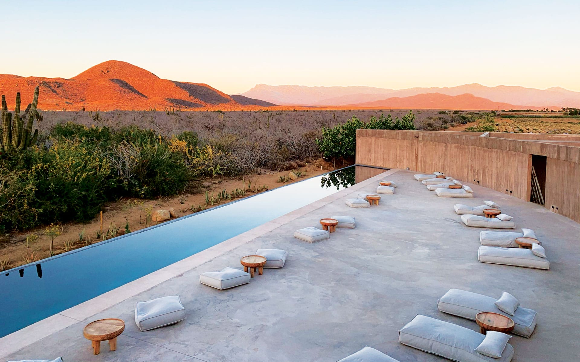 Desert landscape with a modern outdoor seating area and infinity pool at sunset, featuring mountains and cacti in the background.
