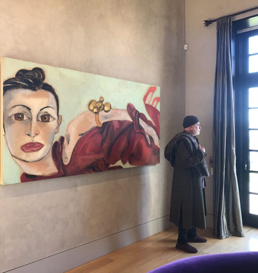 Man in coat and hat stands by a large painting of a woman with striking eyes and red clothing, near a sunlit window.