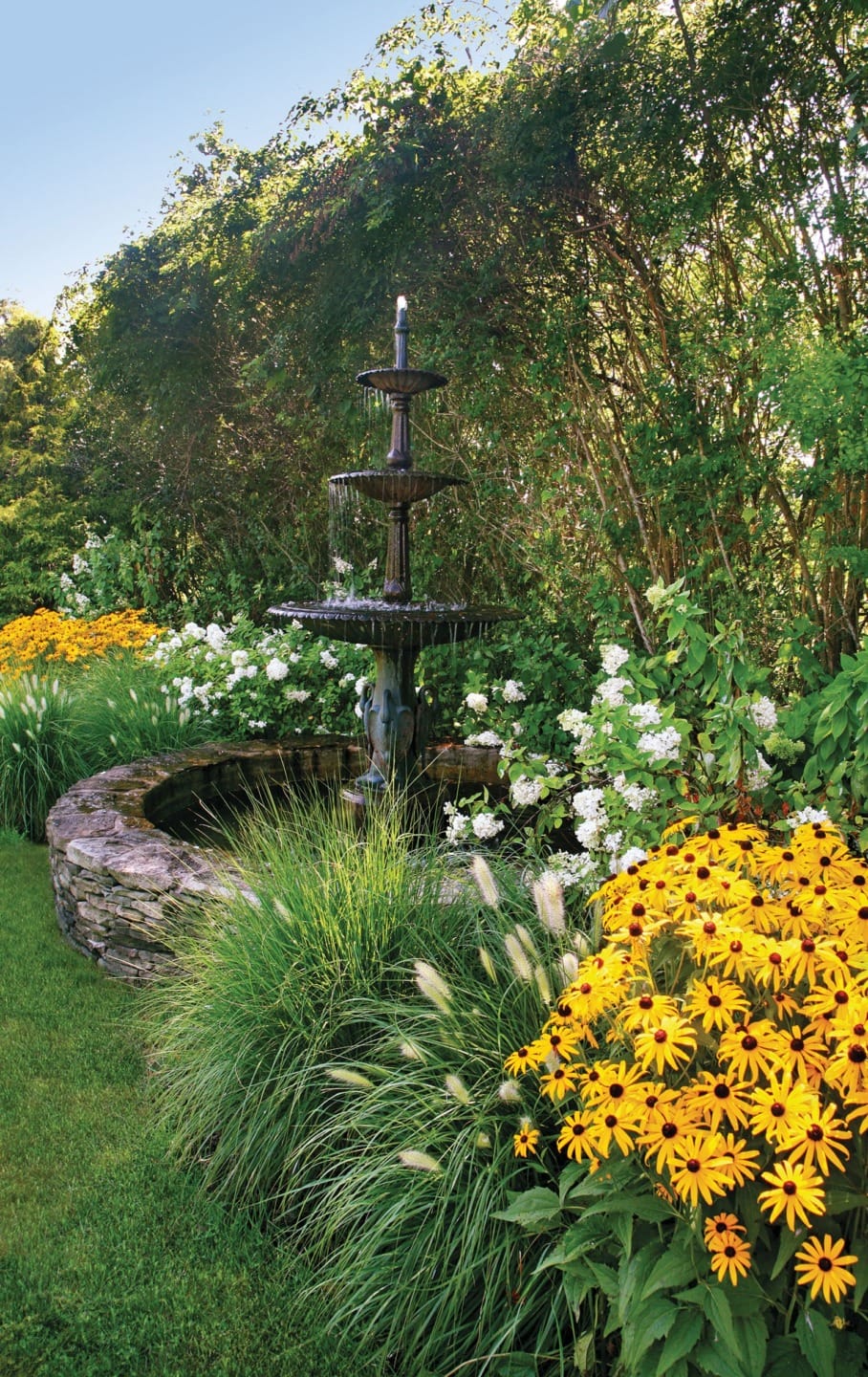 Garden scene with a three-tiered fountain surrounded by yellow and white flowers and lush greenery.