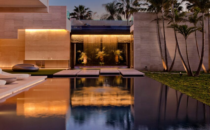Modern luxury home exterior with pool, illuminated waterfall, palm trees, and evening ambiance.