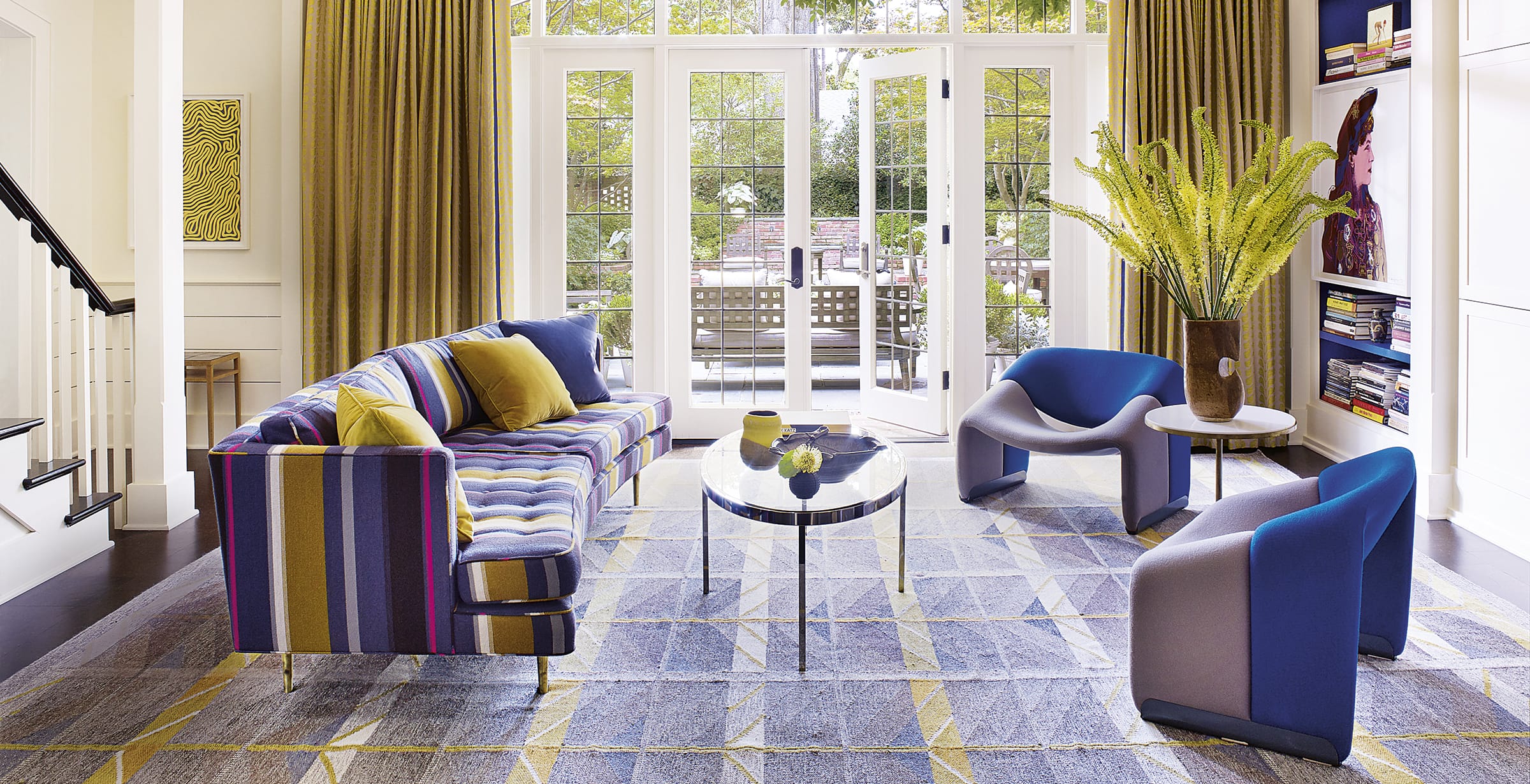 Colorful living room with striped sofa, blue chairs, glass table, and large window with garden view.