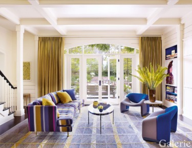 Modern living room with colorful furniture, large windows, yellow curtains, and a geometric rug.